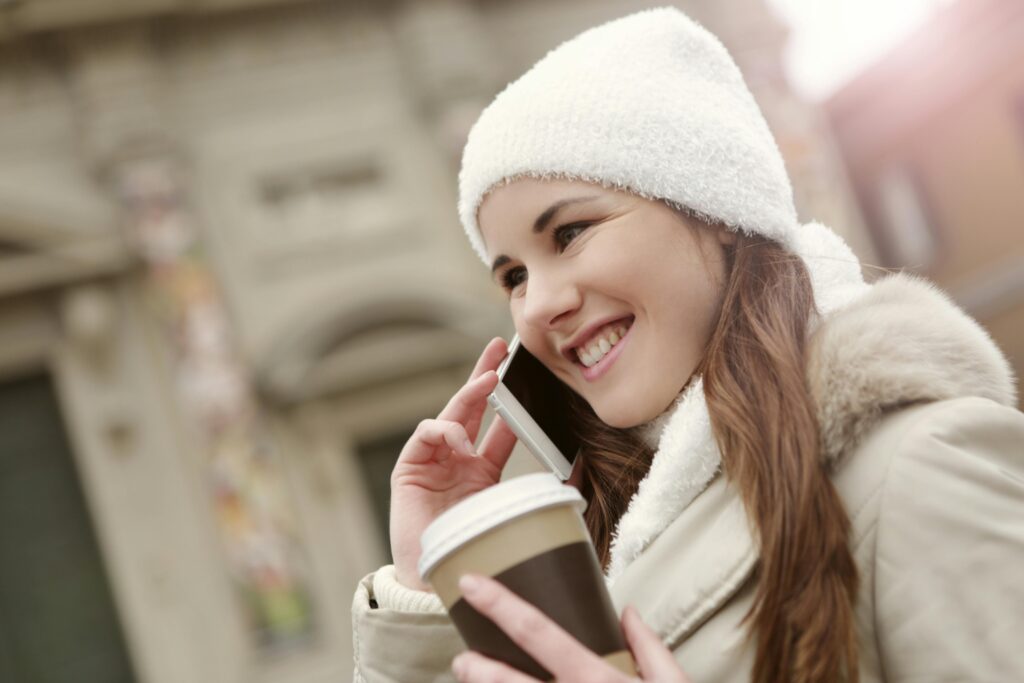 Side view of content female in warm clothes standing in street with hot drink to go and chatting on cellphone while smiling and looking away by Andrea Piacquadio