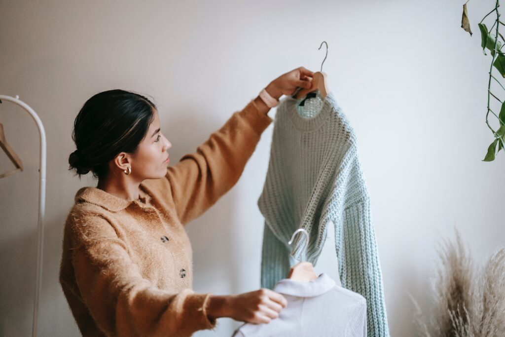 Side view of young pensive female in trendy cardigan looking at knitted sweater and shirt on hangers in cloakroom by Liza Summer