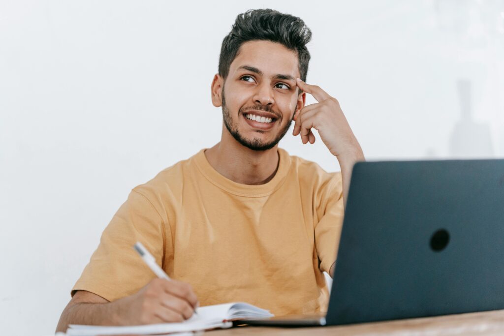 Smiling young bearded Hispanic male entrepreneur thinking over new ideas for startup project and looking away dreamily while working at table with laptop and taking notes in notebook by Michael Burrows