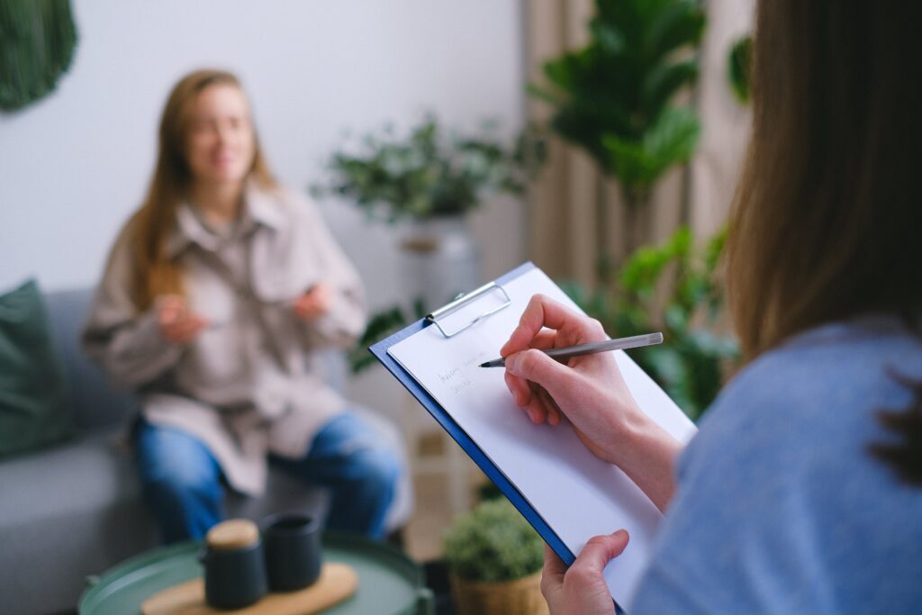 Unrecognizable professional female psychologist writing on clipboard while sitting against client on blurred background during psychotherapy session in light office by SHVETS production