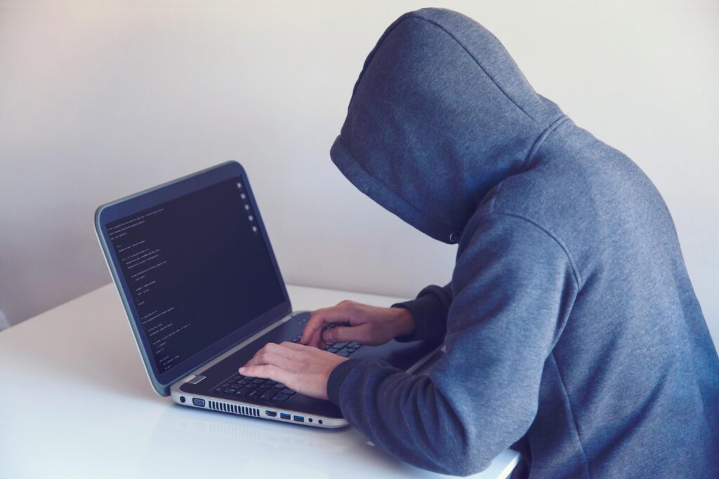 Side view of unrecognizable hacker in hoodie sitting at white table and working remotely on netbook in light room near wall by Nikita Belokhonov