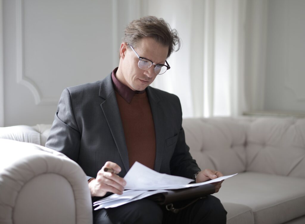 Elegant adult man in jacket and glasses looking through documents while sitting on white sofa in luxury room by Andrea Piacquadio